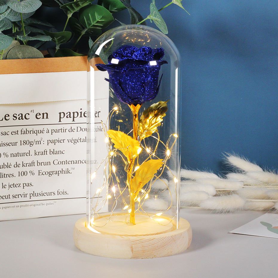 Blue rose and gold leaves in a glass dome with LED lights on a wooden base, against a blue wall with text and plants.