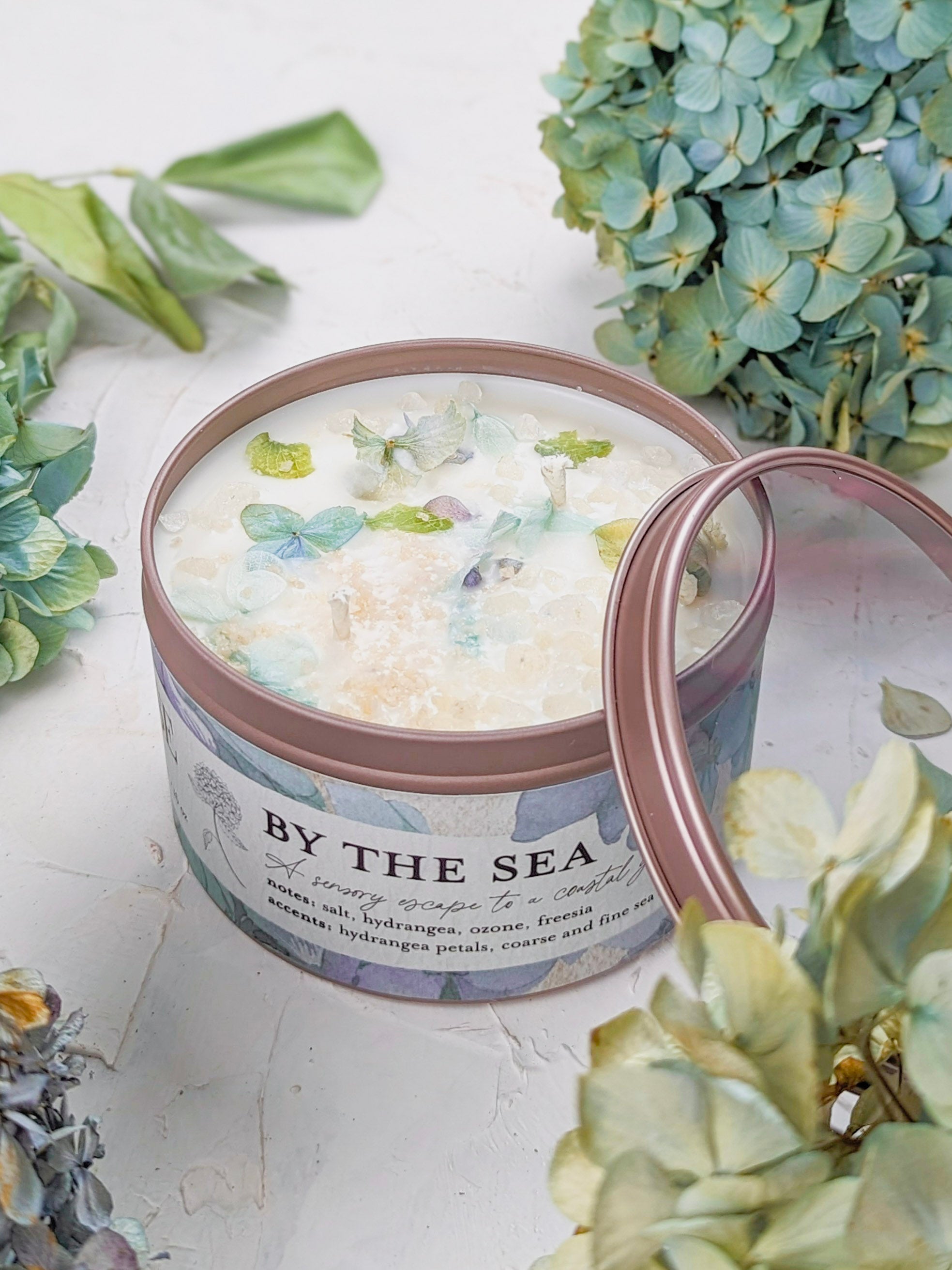 Candle in a tin labeled 'By the Sea' surrounded by floral decorations on a light background