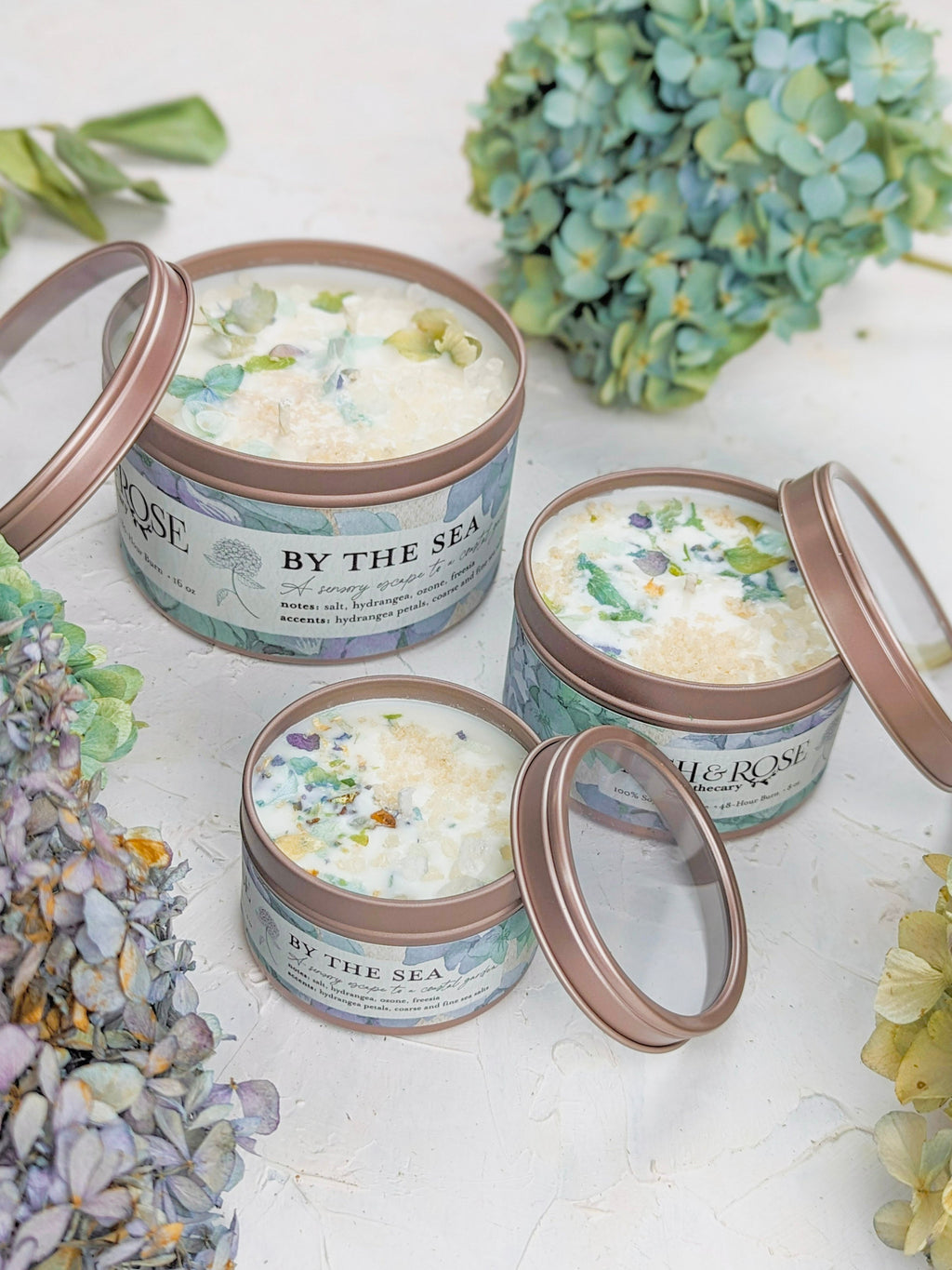 Three tins of 'By the Sea' scented candles with floral designs on a white surface with flowers around.
