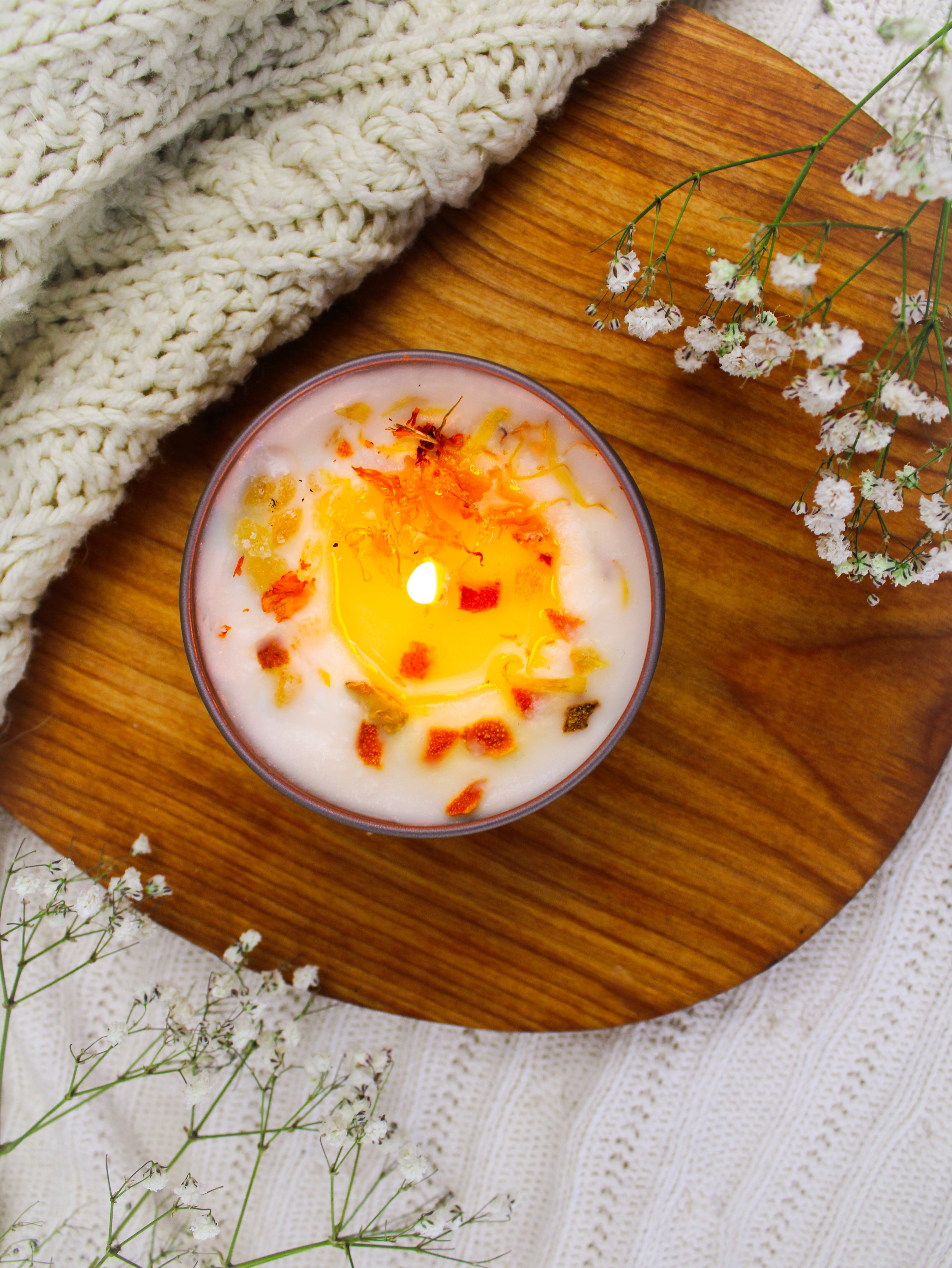 Candle with decorative elements on a wooden board with flowers and fabric in the background