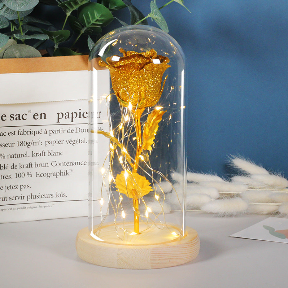 Gold rose in a glass dome with LED lights on a wooden base, placed on a surface with a book and plants in the background.