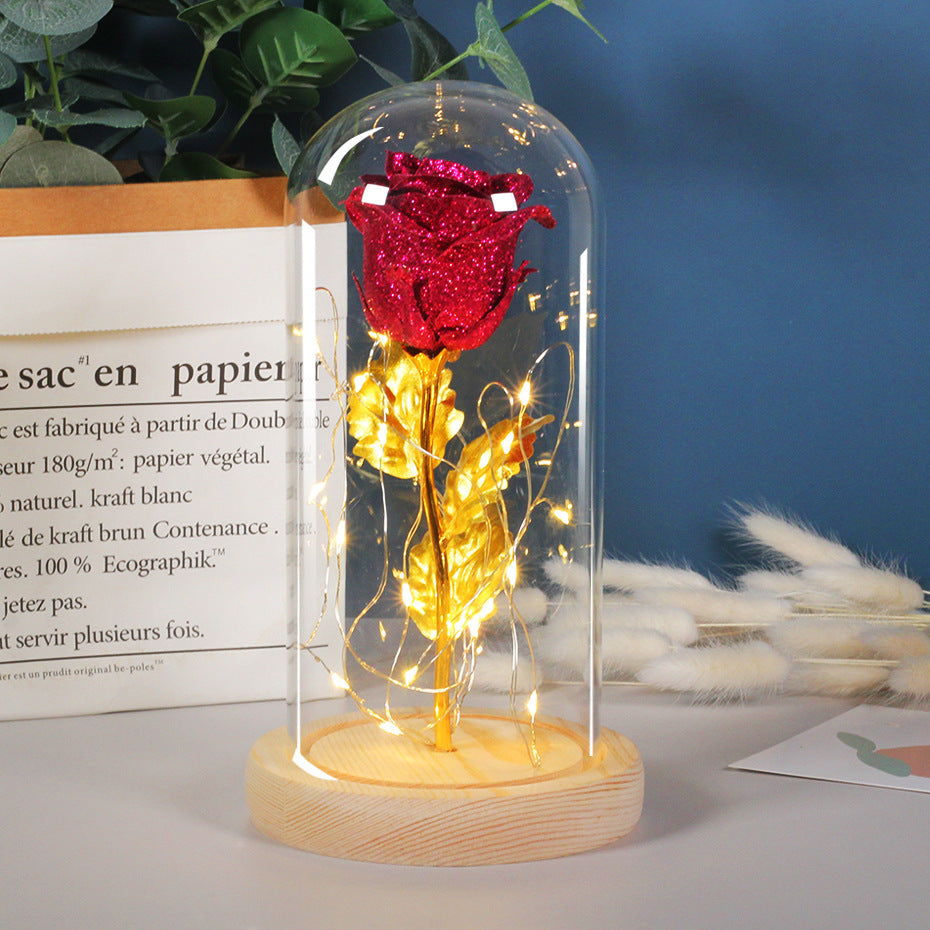 Red rose with gold leaves under a glass dome on a wooden base, surrounded by plants and text.