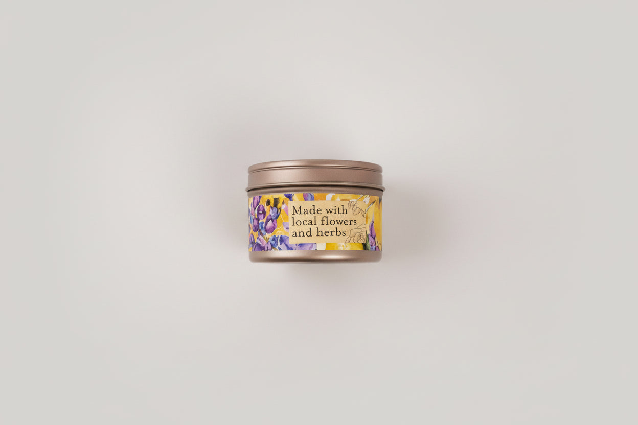 Jar of honey, a tin labeled 'Made with local flowers and herbs', lemons, and purple flowers on a light background