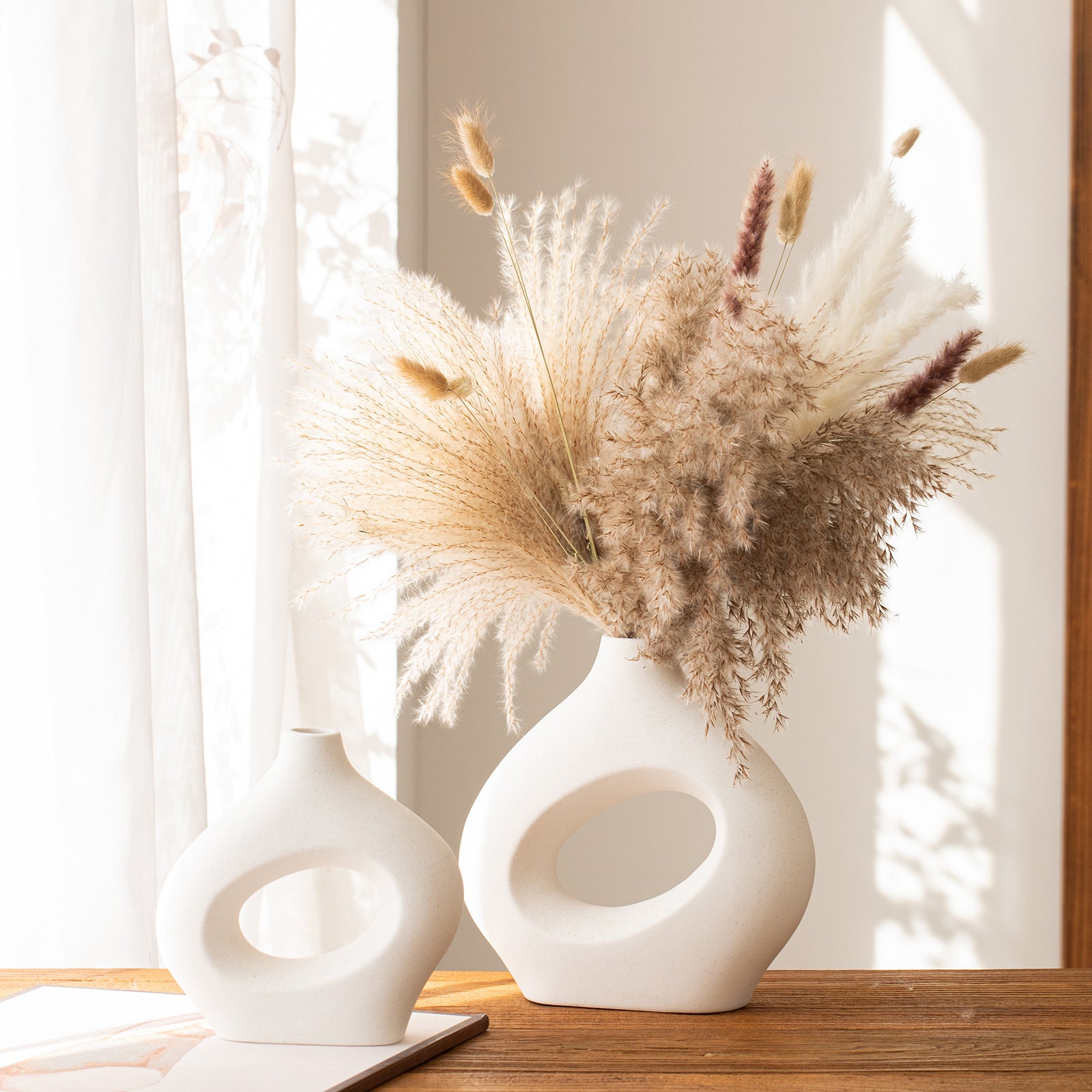 Two white abstract vases with dried pampas grass on a wooden surface.