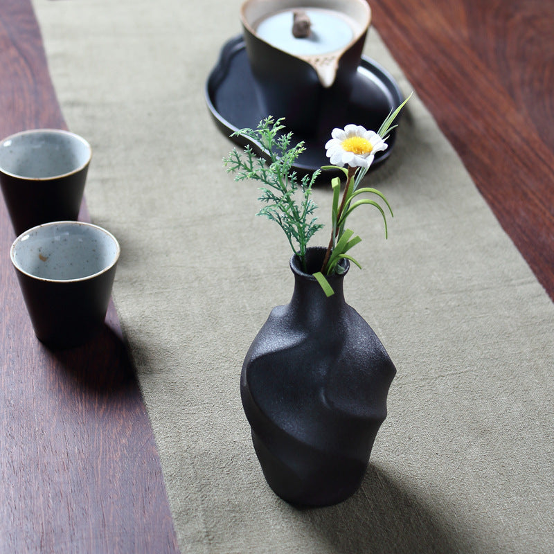 Black vase with flowers on a table with two black cups and a black dish in the background.