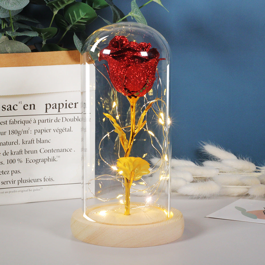 Red and gold rose in a glass dome with LED lights on a wooden base, against a blue wall with plants and text.