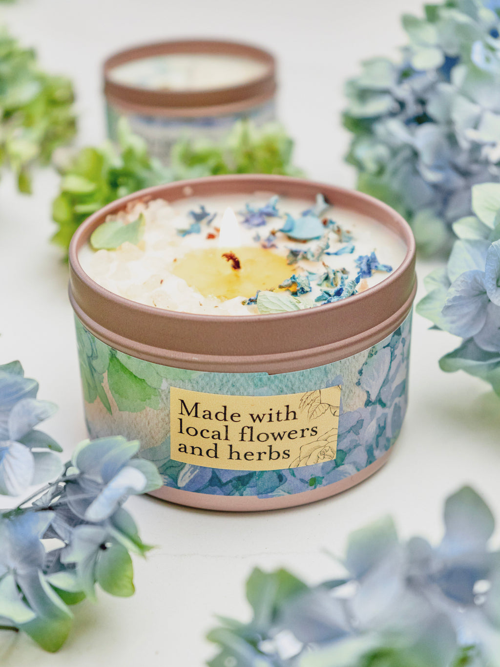 Candle in a decorative tin with floral design and text 'Made with local flowers and herbs' surrounded by flowers.