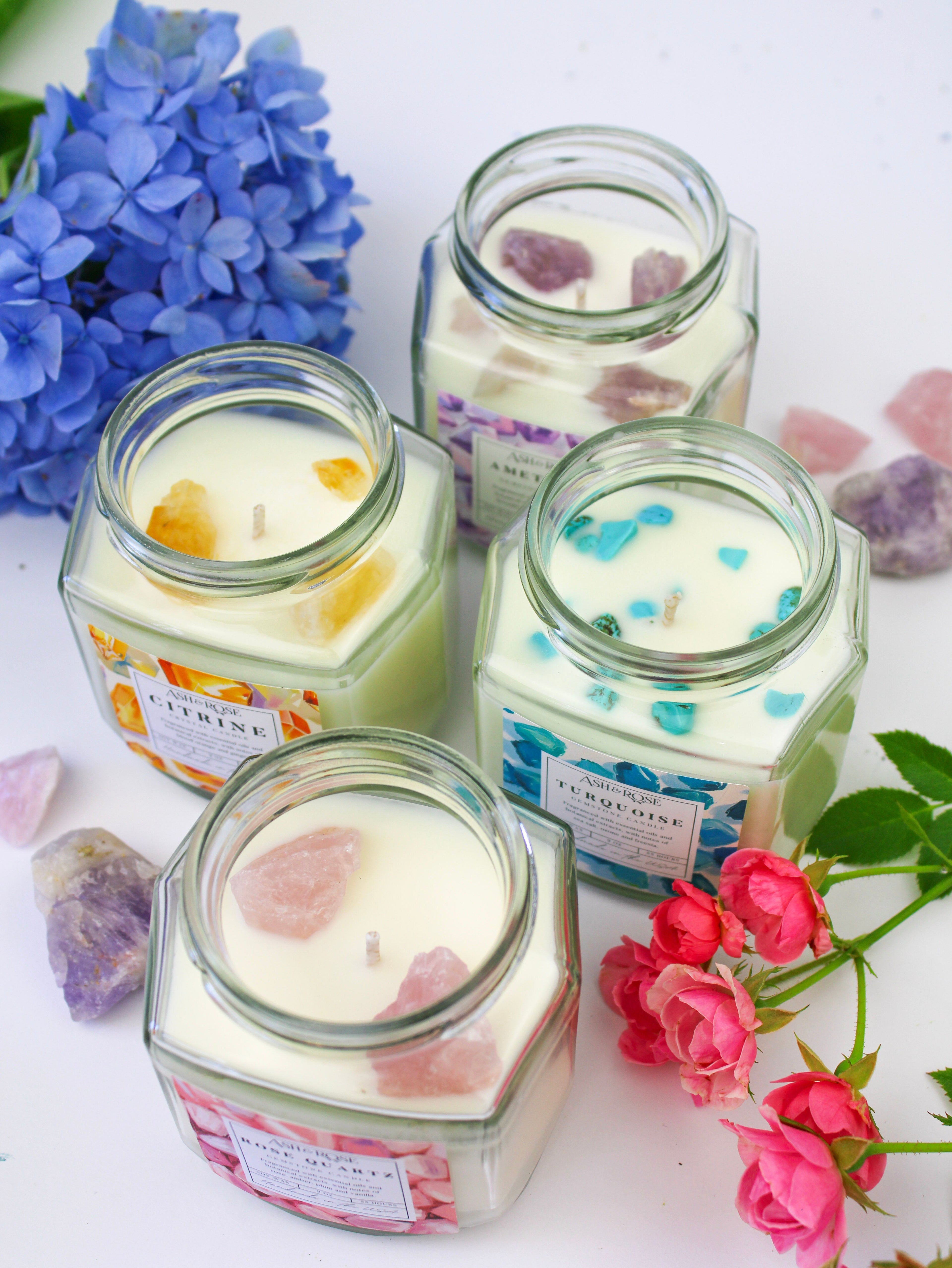 Four hexagonal glass jars with candles labeled 'Citrus' and 'Turquoise' surrounded by flowers on a white background.
