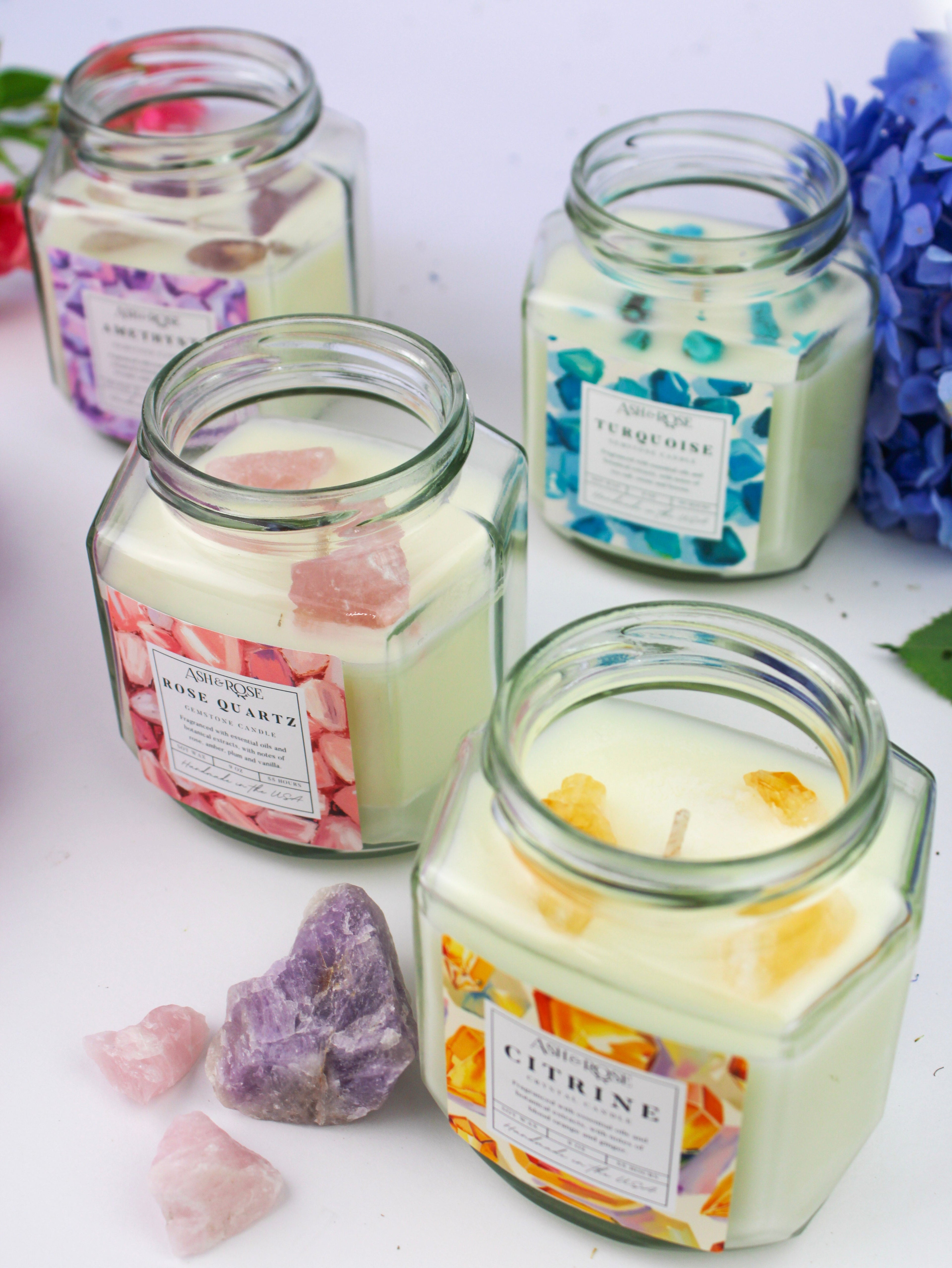 Four glass jars with candles labeled 'Rose Quartz', 'Amber', 'Citrine', and 'Purple Crystal' on a white surface with flowers and crystals.