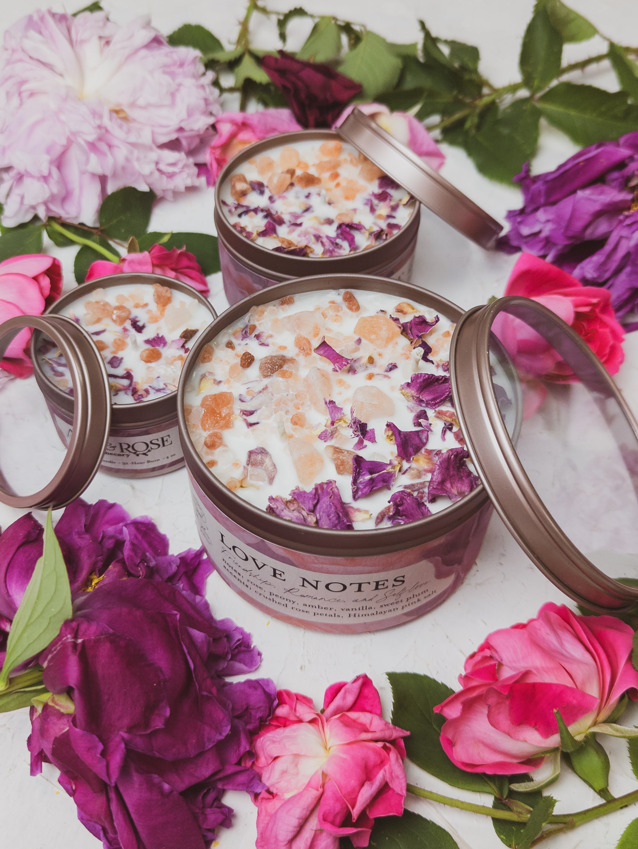 Candle tins labeled 'Love Notes' surrounded by pink and purple flowers on a white surface