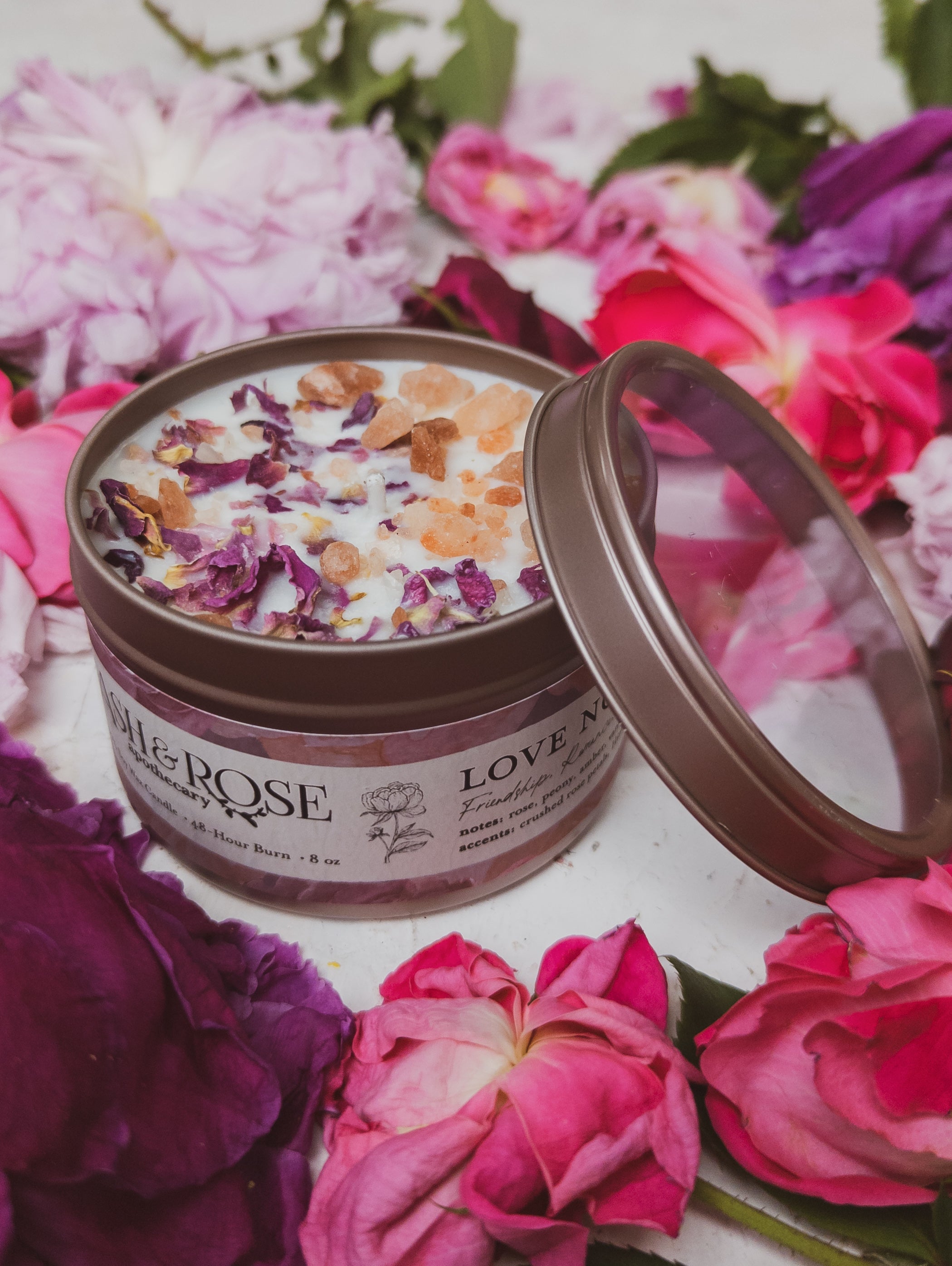 Candle in a tin with rose petals surrounded by pink and purple flowers