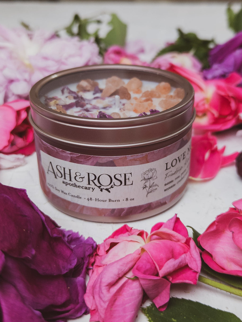 Candle in a tin with 'Ash & Rose Apothecary' branding surrounded by flowers