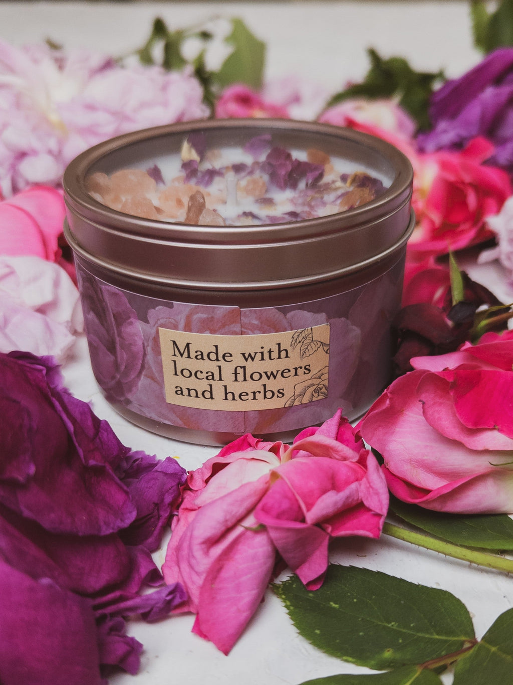 Candle in a tin surrounded by pink and purple flowers with a label indicating local ingredients.
