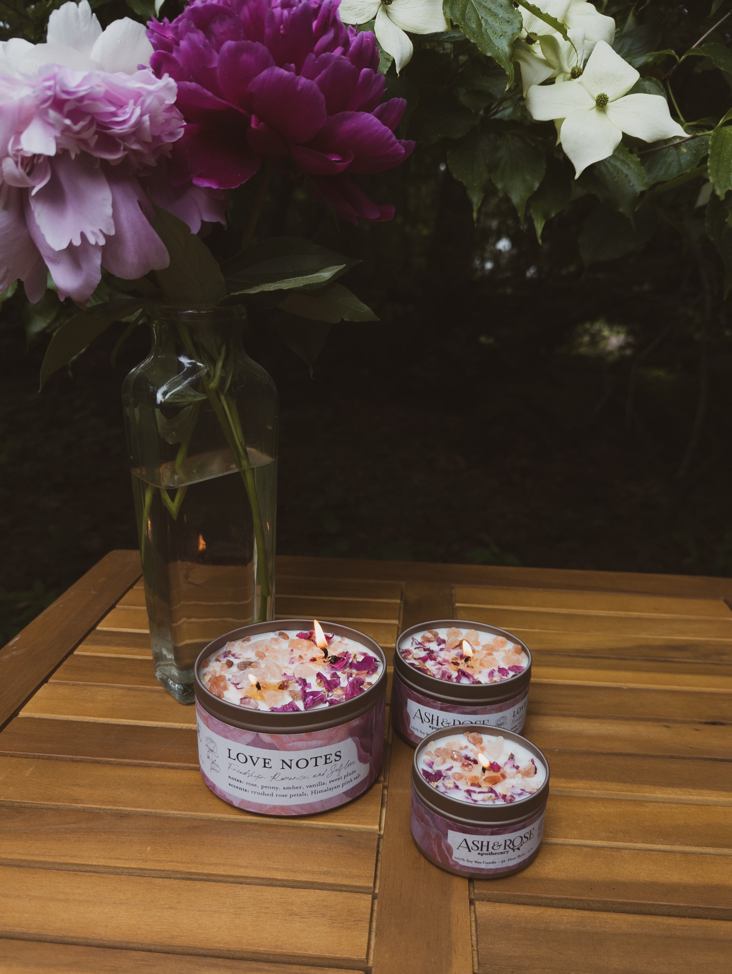 Three candles labeled 'Love Notes' on a wooden surface with flowers in the background