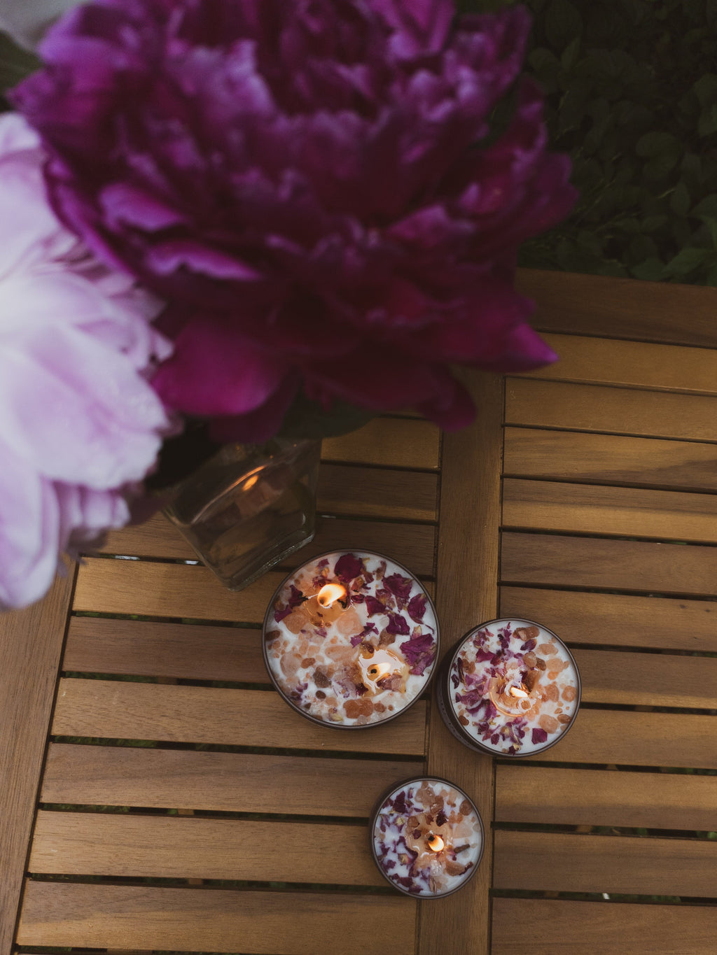 Three small candles with floral designs on a wooden surface with pink and purple flowers in the background.
