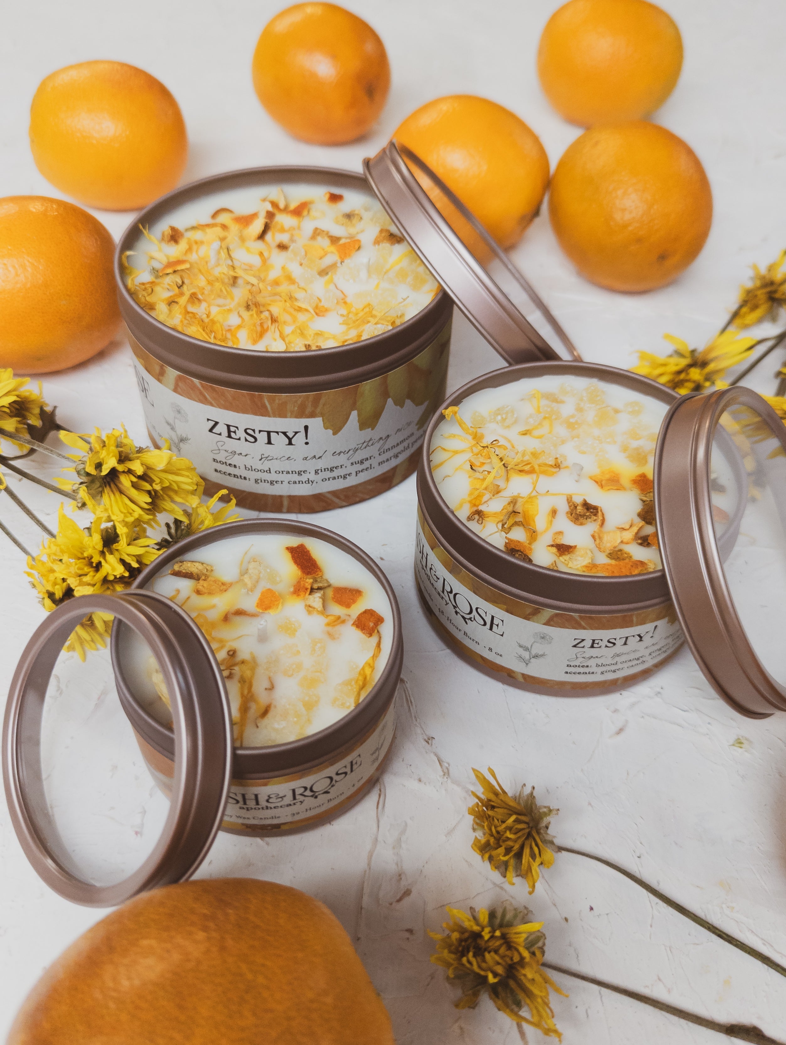 Three tins of 'Zesty!' body scrub with oranges and flowers on a white background