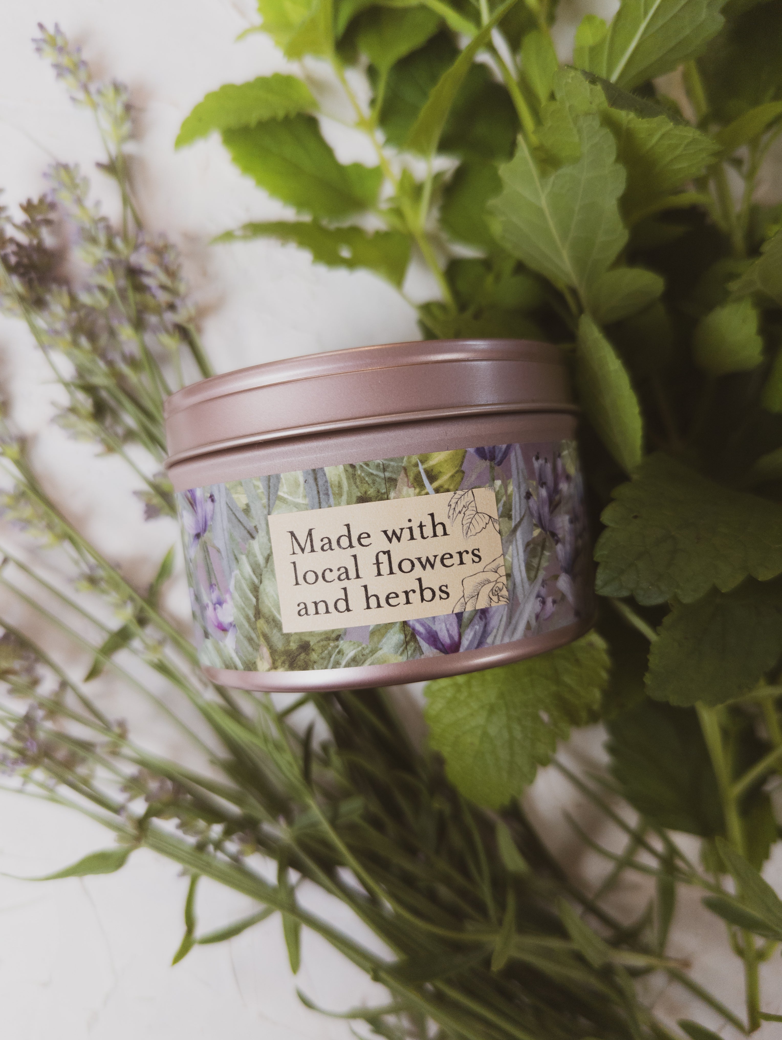 Pink tin labeled 'Made with local flowers and herbs' surrounded by green leaves and lavender.