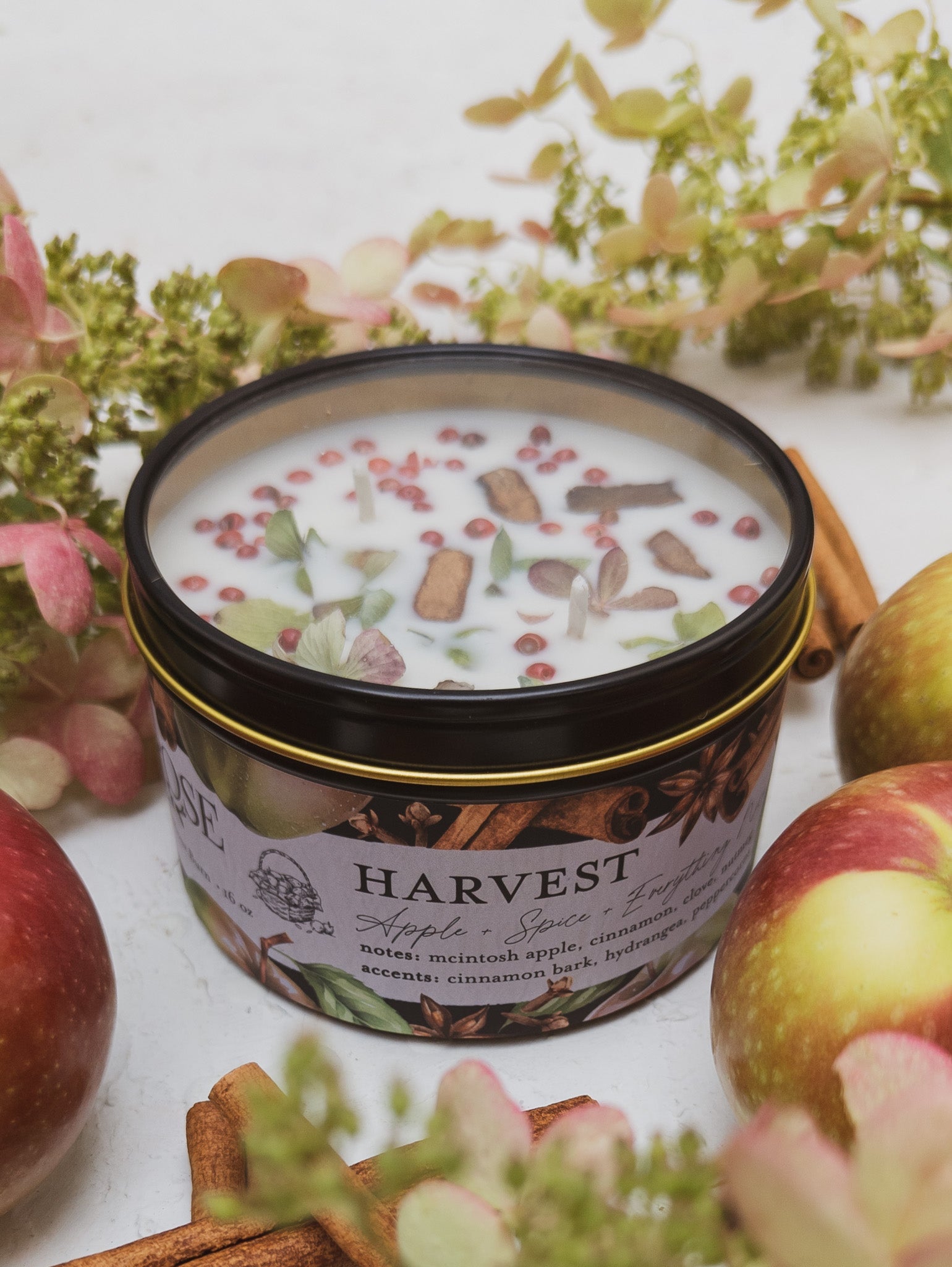 Candle in a tin labeled 'Harvest' surrounded by apples, cinnamon sticks, and floral elements on a white background.