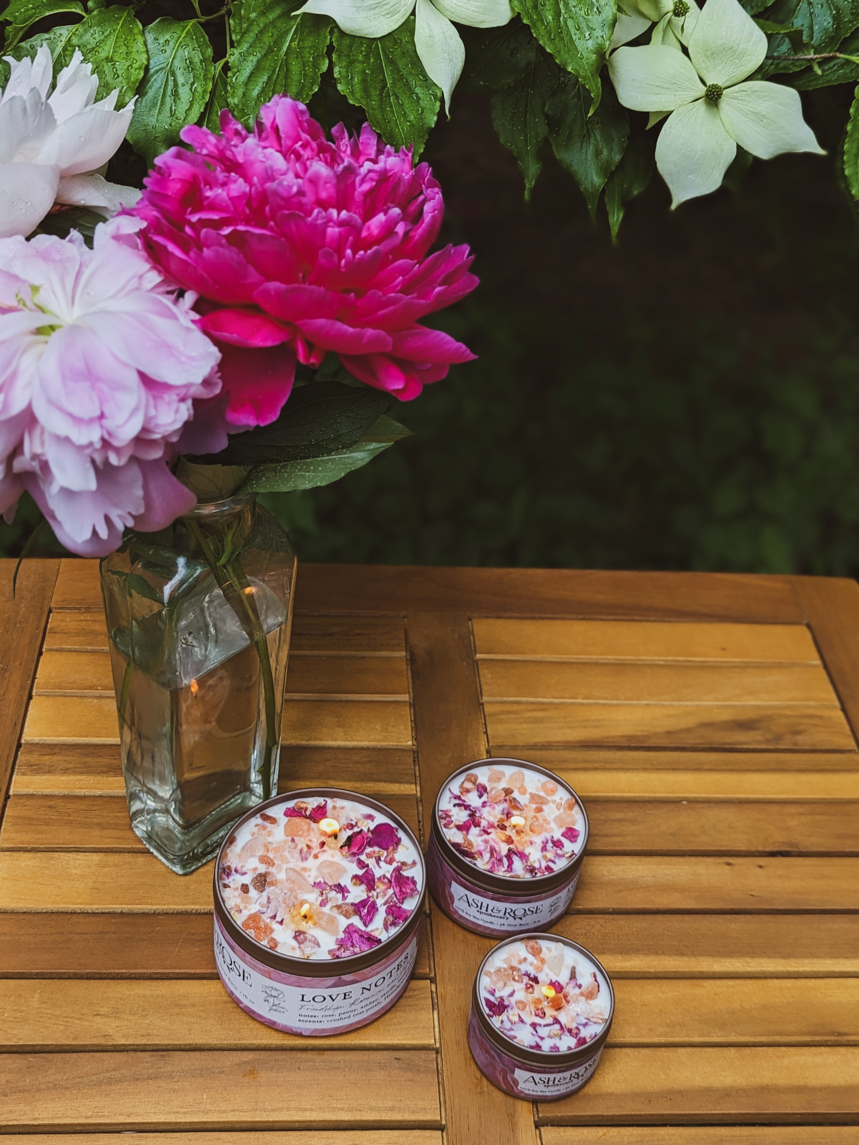 Three small jars with decorative lids on a wooden surface next to a vase of flowers.