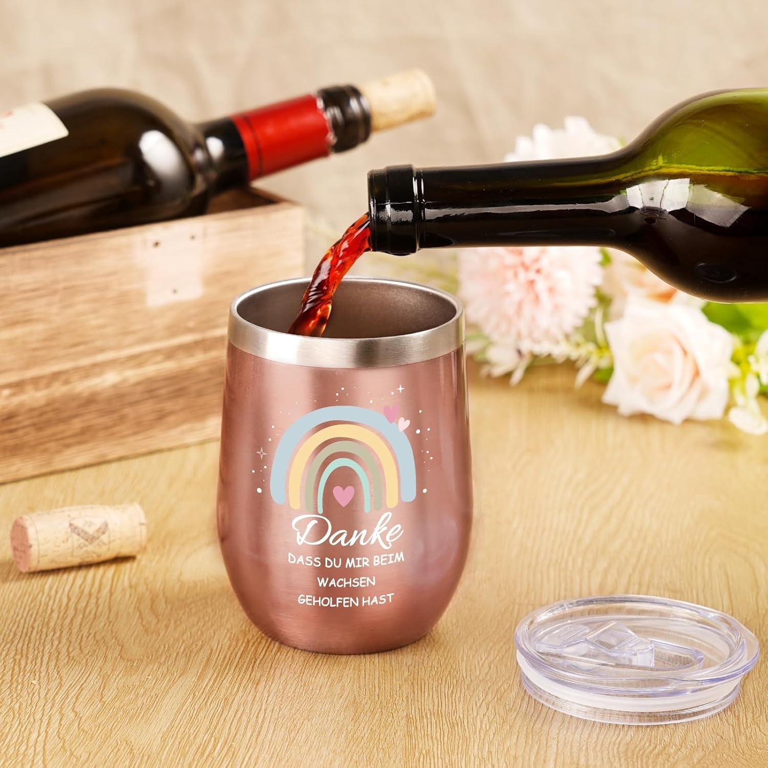 Rose gold wine tumbler with rainbow design and text being filled with red wine on a wooden surface.
