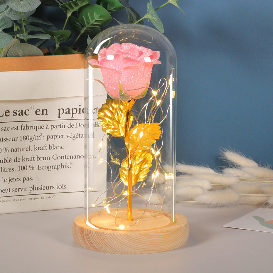 Pink and gold rose in a glass dome with LED lights on a wooden base, placed on a surface with a blurred background.