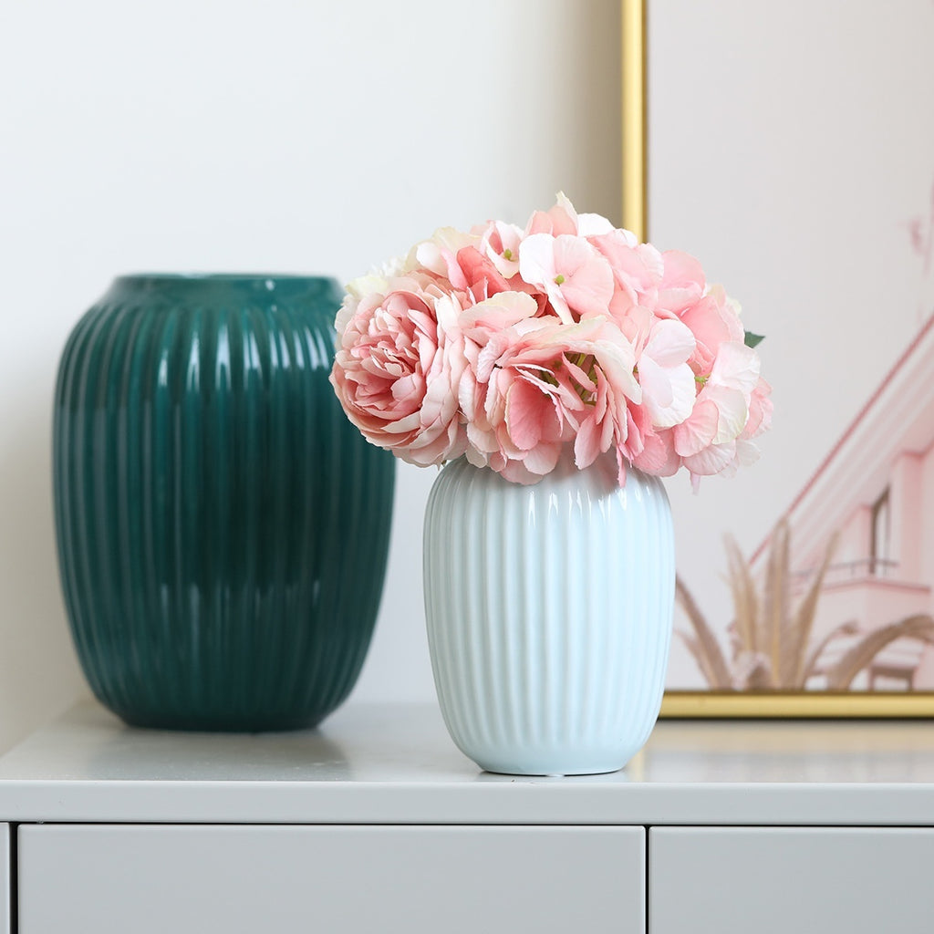 White vase with pink flowers next to a green vase on a surface.